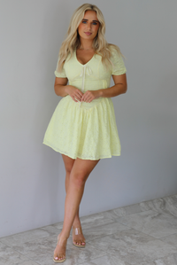Woman wearing a light yellow mini dress with a floral eyelet design throughout, v-neckline, short sleeves, fitted drop waist bodice, and a flowy mini length skirt indoors against a white wall with white marble flooring. 