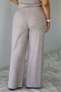 Woman wearing a taupe colored two piece set featuring a vest-appearance top with non-functional button closures on the left side and high waisted pants. The photo shows a closeup of the back of the bottoms. The photo is taken indoors against a white wall with white marble flooring. 