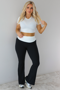 Woman wearing a two piece set featuring a white cropped top with a black polka dot print pattern and black pants that are high waisted with a white and black polka dot print folded over detail. The photo is taken indoors against a white wall with white marble flooring.