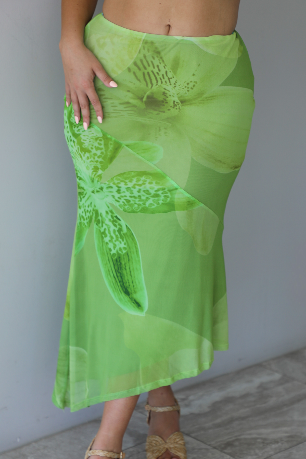 Woman wearing a green tropical print set featuring a backless tank and high waisted, asymmetrical hem midi skirt indoors against a white wall with white marble flooring. The photo shows a closeup of the front of the skirt. 