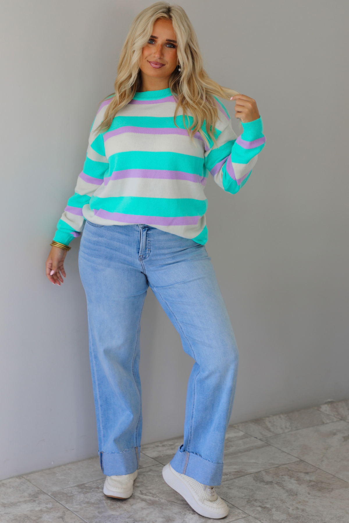 A woman wearing a turquoise, lavender, and cream striped knit sweater, paired with light wash denim jeans, standing indoors against a white wall with white marble flooring. 
