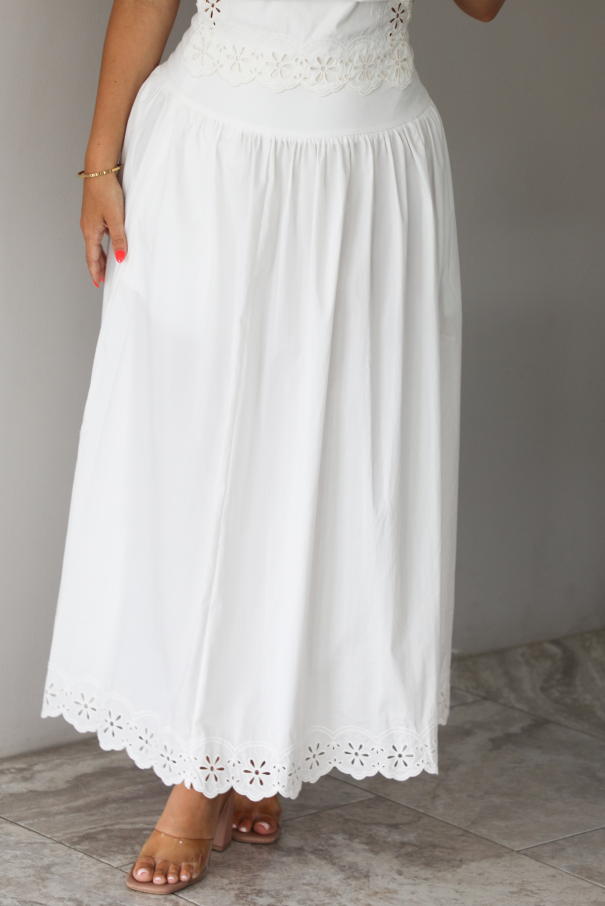 Woman wearing a two piece white set indoors against a white wall with white marble flooring. The photo shows a closeup of the skirt, and shows how it has eyelet details at the hem. 
