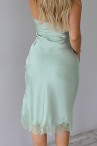 Woman wearing a light sage green, satin midi dress that has a strapless design and a split hem detail with a lace trim indoors against a white wall. The photo shows the back of the dress. 