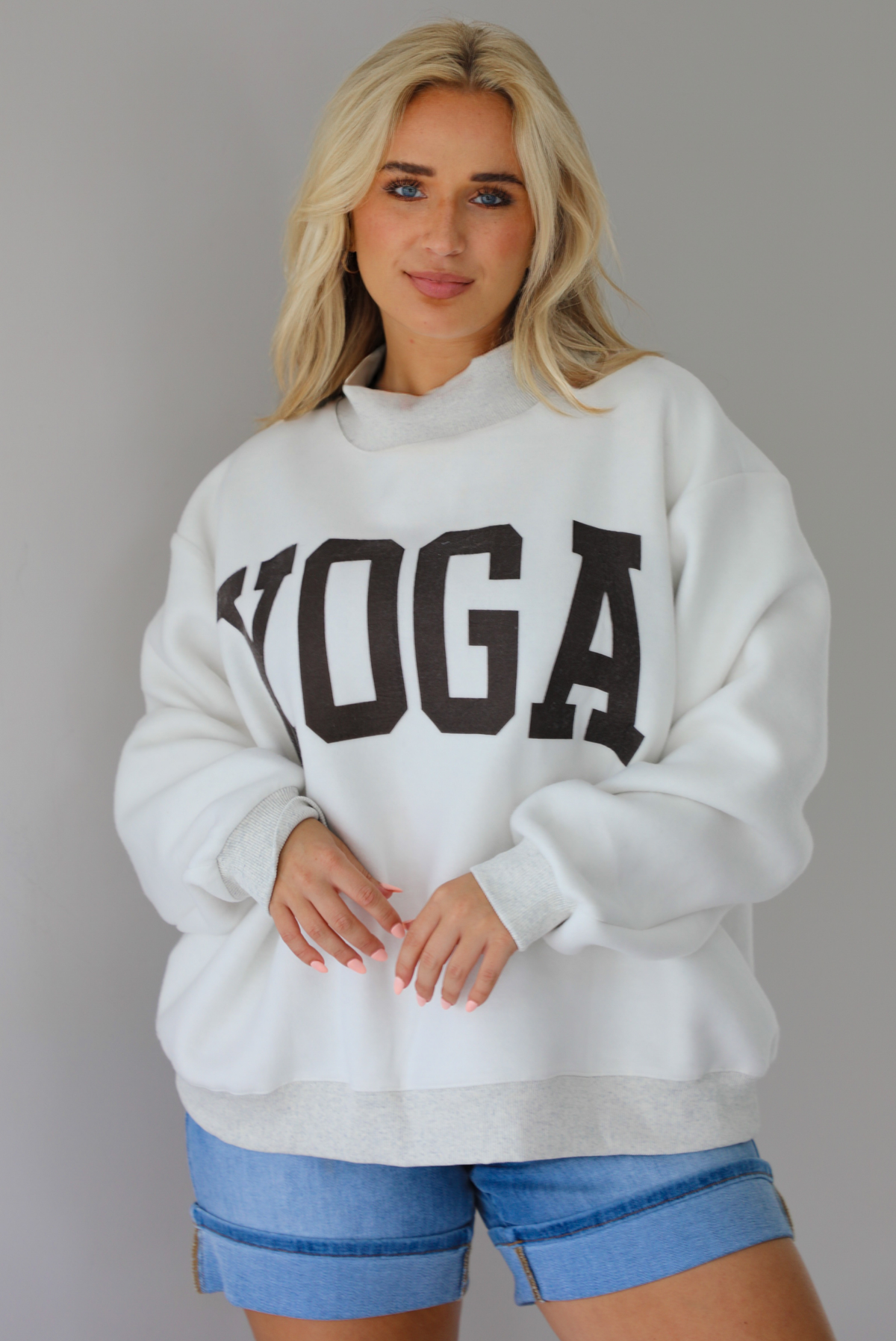 Woman wearing a heather grey oversized sweatshirt, reversed inside out, with the word 'yoga' written in collegiate font on the front with medium wash denim shorts indoors against a white wall.