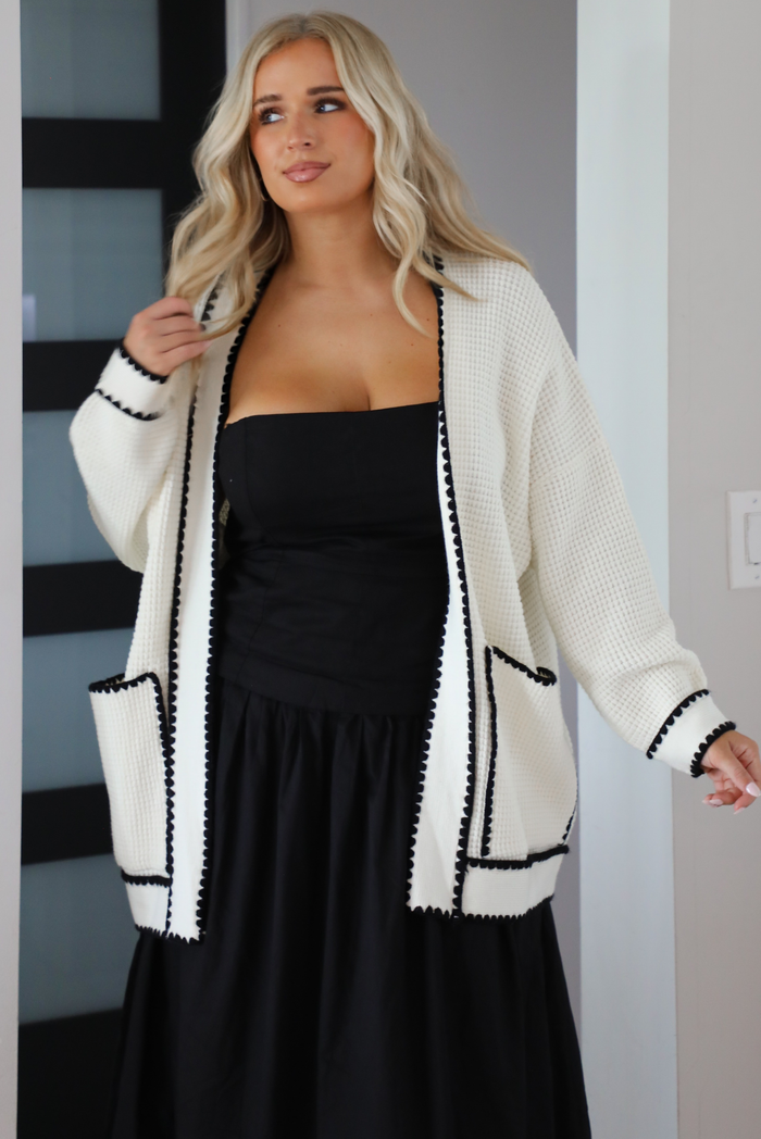 Woman model wearing a cream colored oversized cardigan that has black trim details along the inner hems, at the wrists, and at the pockets. The cardigan is worn with a black strapless midi dress underneath. The photo is taken indoors a modern home with a white and black interior. 
