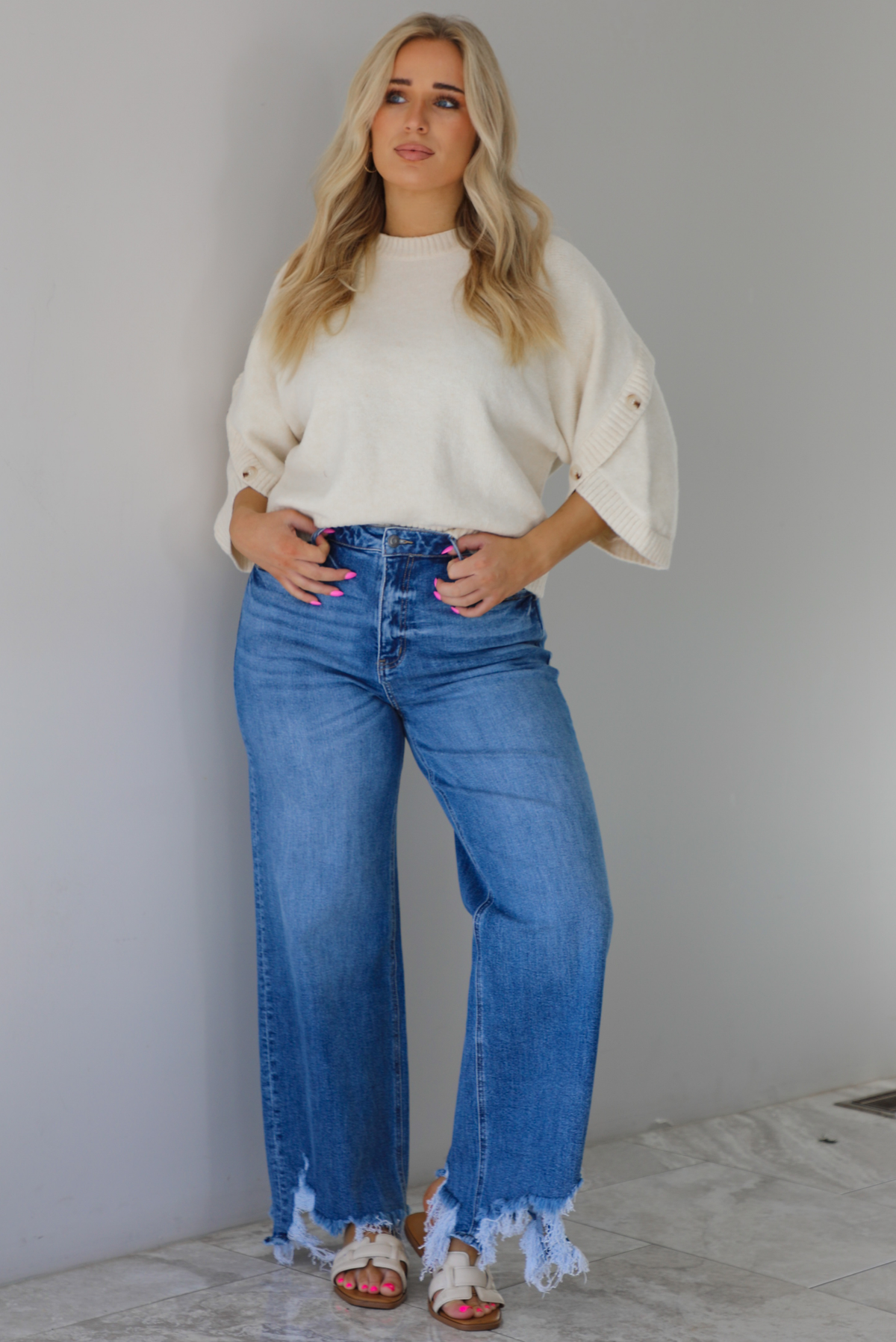 Woman wearing a cream colored quarter sleeve sweater that has ribbed details at the neckline, on the sleeves, and on the hem, with non-functional button closures on the sleeves. The sweater has a relaxed fit and is paired with a medium wash, high waisted, distressed hem denim with sand colored sandals. The photo is taken indoors against a white wall with white marble flooring. 