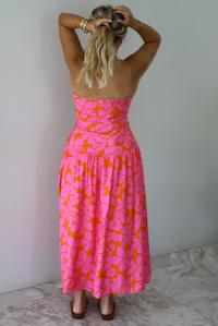 A woman viewed from behind wearing a strapless pink and orange floral print maxi pattern, standing indoors against a white wall with white marble flooring. The back of the dress has a zipper closures. 