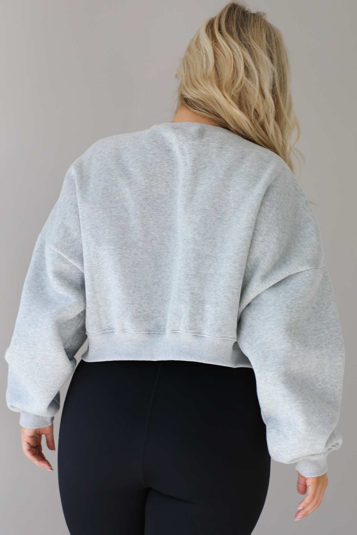 Woman wearing a heather grey cropped sweatshirt with a black fitted jumpsuit underneath. The photo shows a closeup of the back of the sweater. The photo is taken indoors against a white wall.