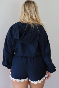 A woman shown from behind modeling a matching navy blue two-piece set with white floral lace trim, consisting of a cropped zip-up jacket with a hoodie detail and shorts, standing indoors against a white wall.