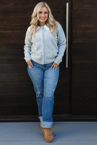 Woman model wearing a heather grey cardigan with a white lace bib at the neckline, has functional button closures in the front, functional pockets in the front, and a relaxed fit. The cardigan is paired with a medium wash denim and tan ugg boots. The photo is taken outdoors against a dark brown wooden door.