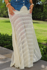 A woman outdoors wearing a long cream crochet lace maxi skirt with a denim top. The background shows a green lawn and trees.