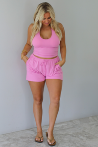 A woman wearing a matching pink activewear set consisting of a halter crop top and drawstring shorts, standing indoors against a white wall with white marble flooring.