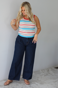 A woman modeling a sleeveless ribbed knit top with horizontal stripes in red, light blue, navy, and white, paired with dark wide-leg jeans and metallic rose gold flats, standing indoors against a white wall with white marble flooring. 