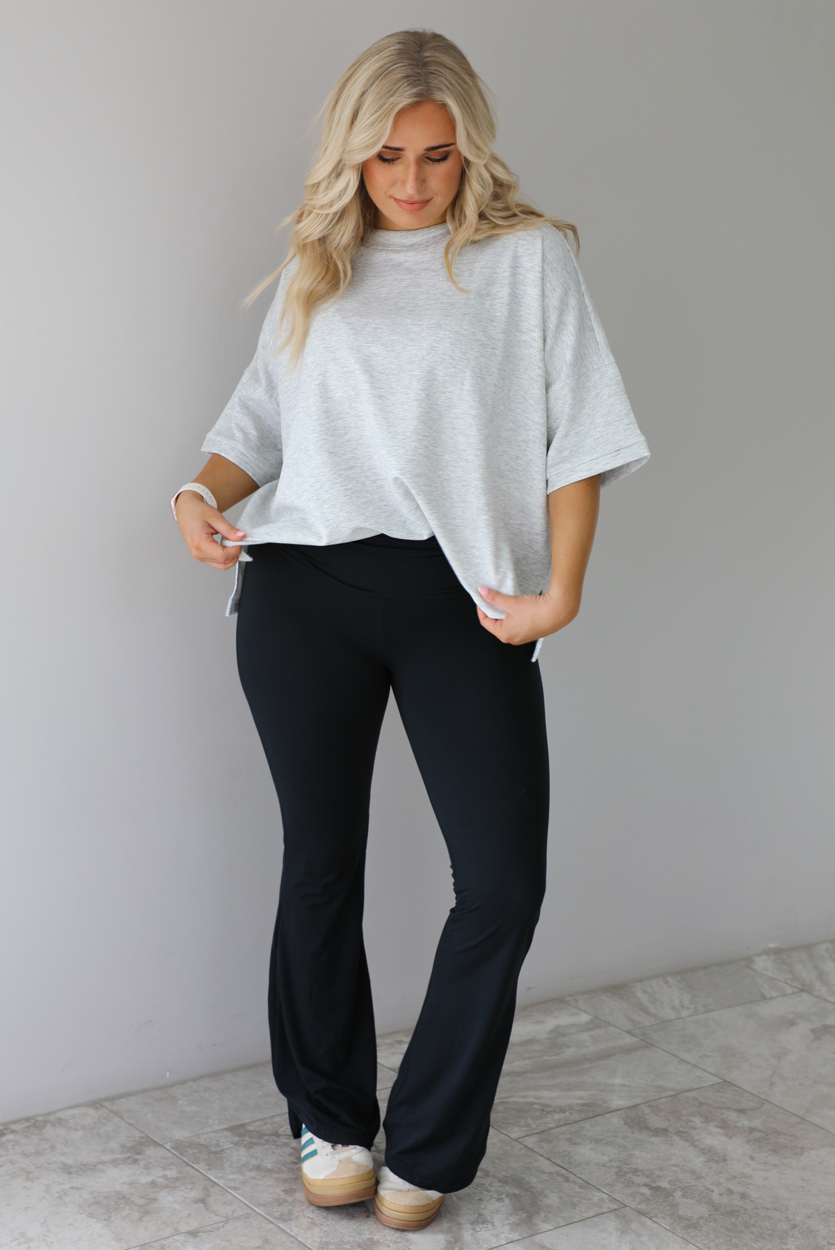 Woman wearing an oversized heather grey box tee with black leggings that flare slightly at the hems. The photo is taken indoors against a white wall with white marble flooring. 