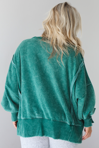 Woman wearing a green colored oversized sweatshirt with exposed seam details and a high-low design with heather grey casual pants. The photo is taken indoors against a white wall. The photo shows the back of the sweater. 
