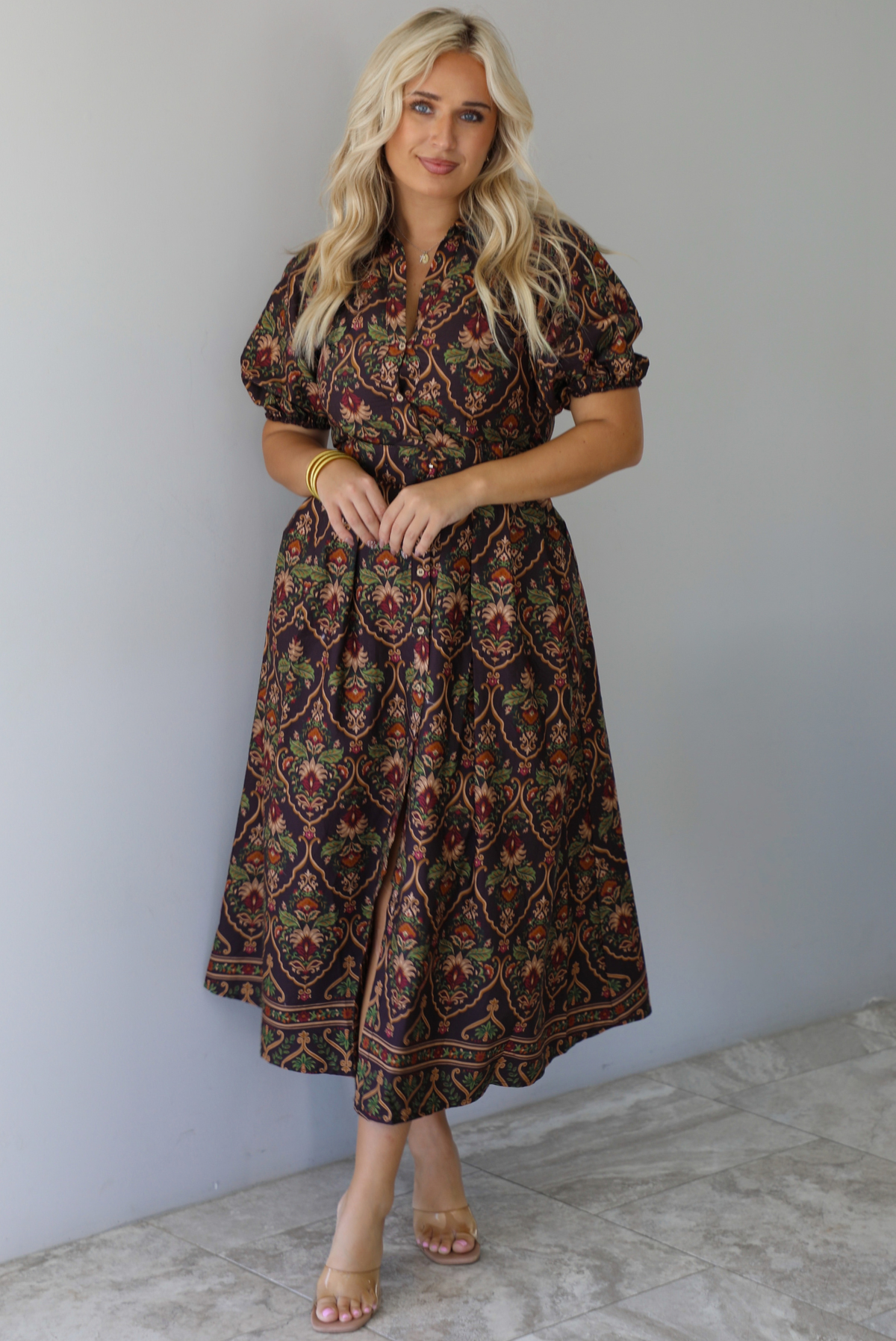 A woman wearing a dark brown midi dress with a multi-colored design throughout, a collared neckline, short puffed sleeves, functional button closures along the front, a belted detail at the waistline, standing indoors against a white wall with white marble flooring. 