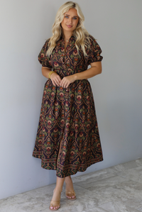 A woman wearing a dark brown midi dress with a multi-colored design throughout, a collared neckline, short puffed sleeves, functional button closures along the front, a belted detail at the waistline, standing indoors against a white wall with white marble flooring. 