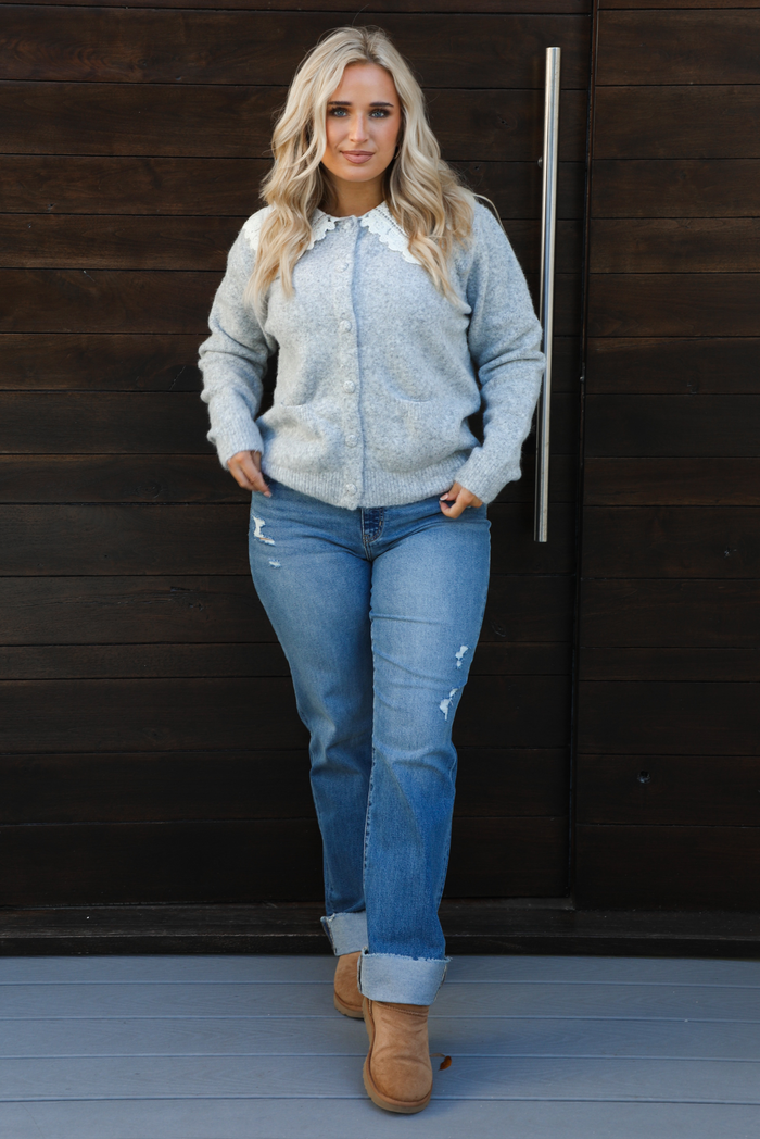 Woman model wearing a heather grey cardigan with a white lace bib at the neckline, has functional button closures in the front, functional pockets in the front, and a relaxed fit. The cardigan is paired with a medium wash denim and tan ugg boots. The photo is taken outdoors against a dark brown wooden door.
