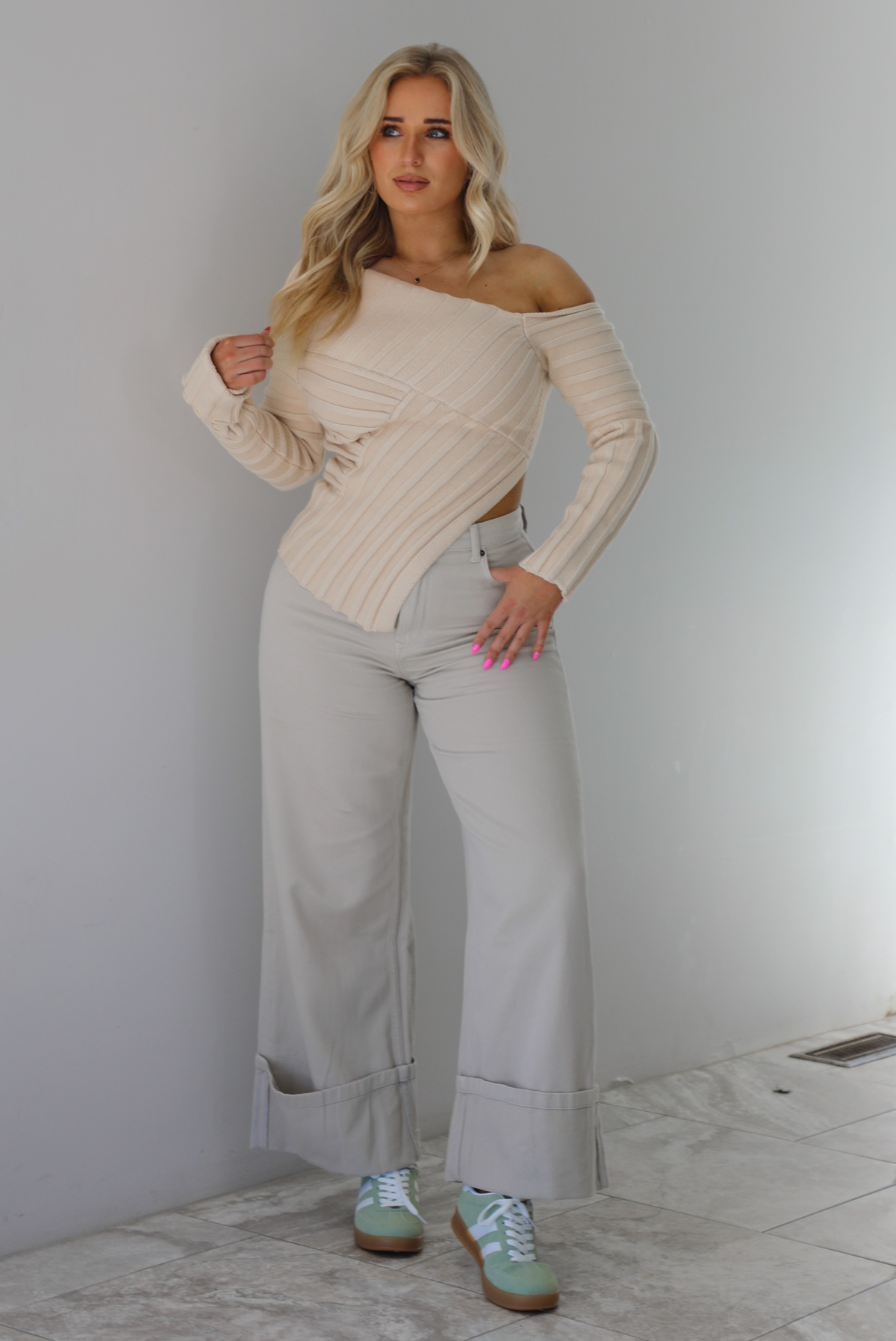 Woman wearing a cream colored ribbed textured knit top that has an asymmetrical hem with long sleeves in which one side is worn off the shoulder. The sweater is paired with sand colored high waisted, cuffed hem denim. The photo is taken indoors against a white wall with white marble flooring. 