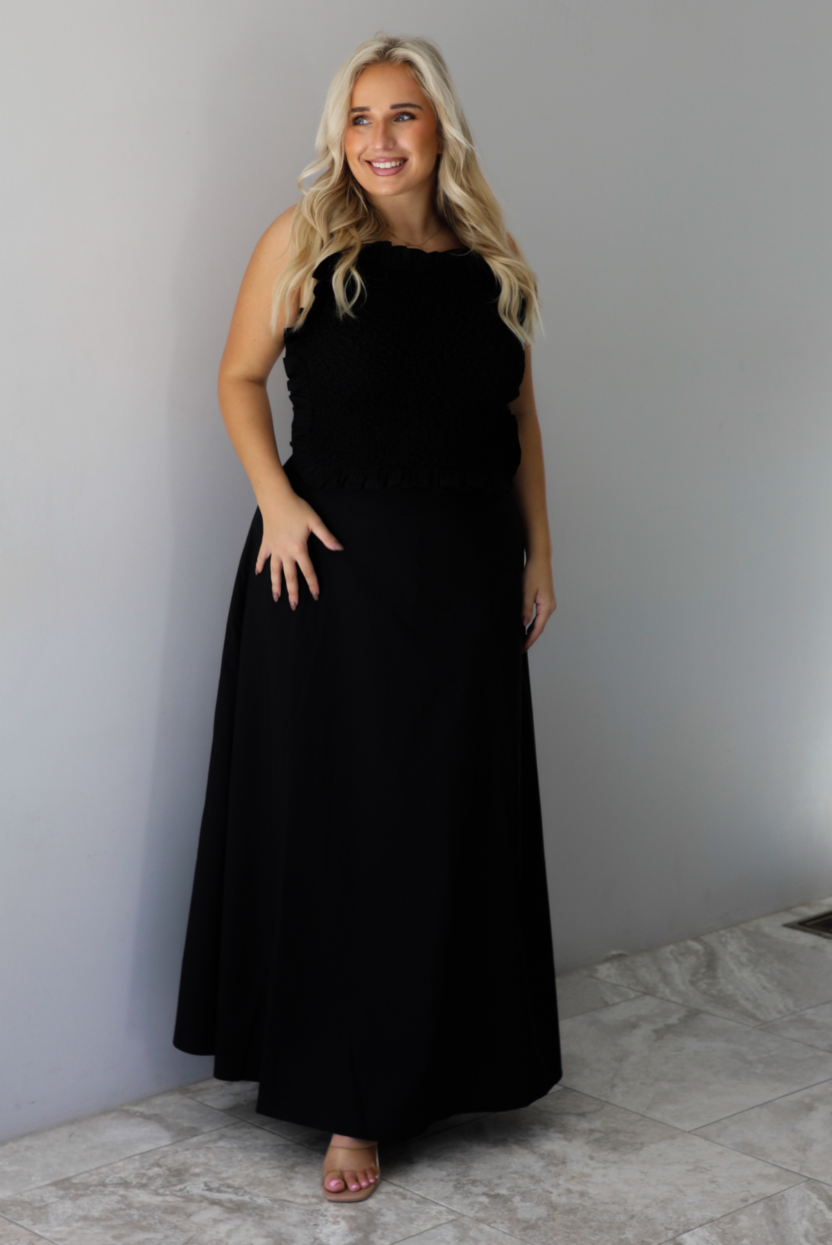 A woman wearing a two piece black set featuring a smocked detail top and high waisted maxi skirt, standing indoors against a white wall with white marble flooring. 