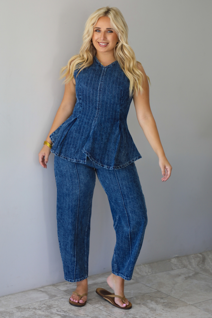 A woman wearing a matching two piece dark wash denim set with a pin striped design on it featuring a sleeveless peplum style top and high waisted denim jeans, standing indoors against a white wall with white marble flooring. 