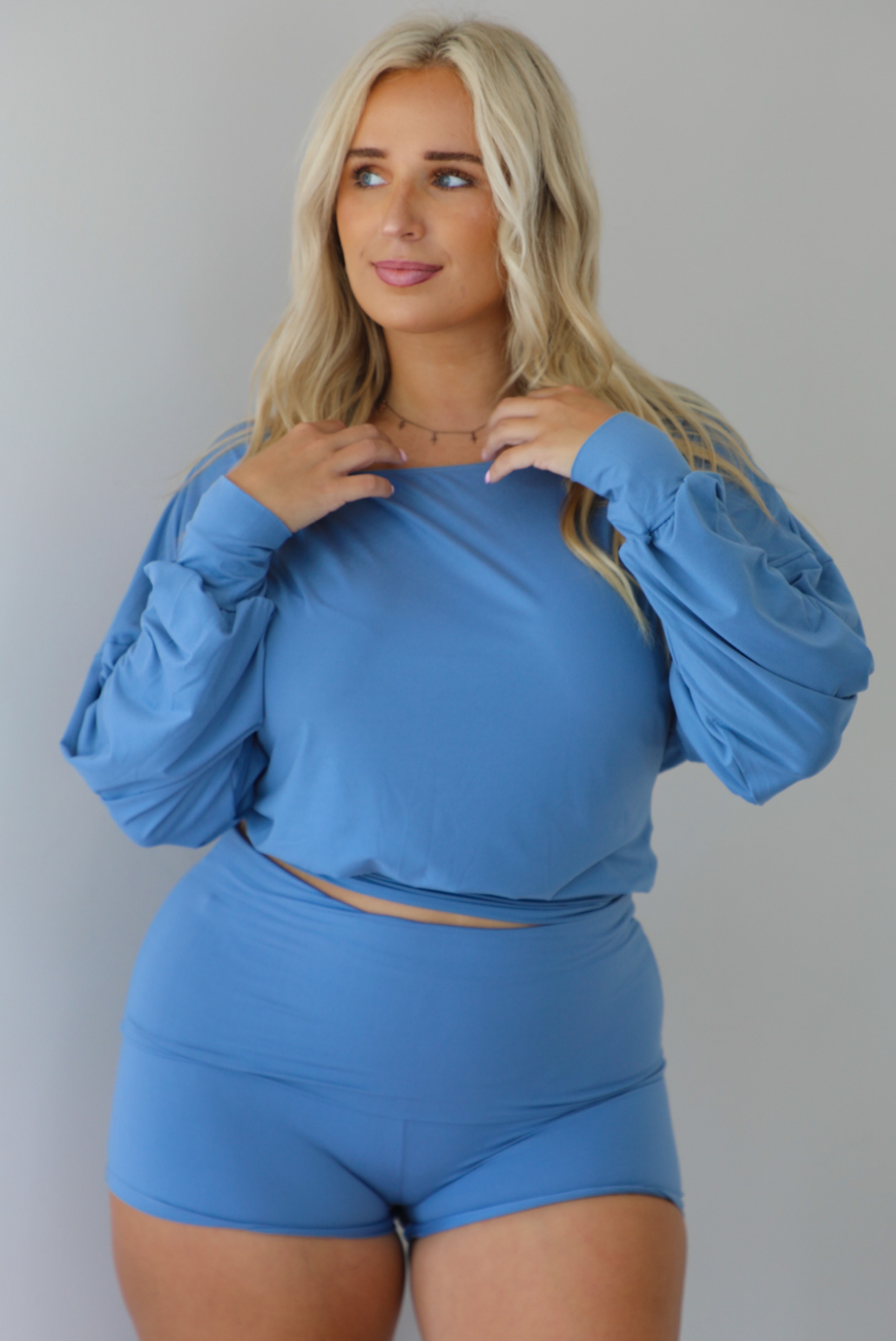 A woman wearing a dust blue matching two piece set consisting of a slouchy off the shoulder top that's fitted at the waist and folded over waist shorts, standing indoors against a white wall.