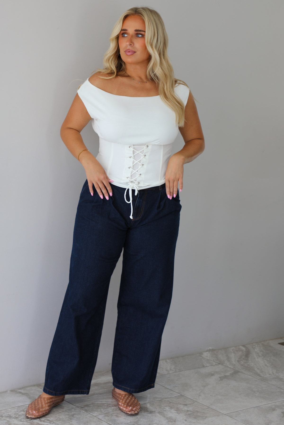 A woman modeling a white off-the-shoulder lace-up corset top paired with dark denim wide-leg jeans and pinkish-brown flat shoes, standing indoors against a white wall with white marble flooring. 