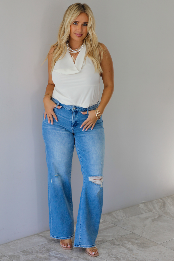 A woman modeling a cream sleeveless halter top and light-wash blue distressed flare jeans, standing indoors against a white wall with white marble flooring. 