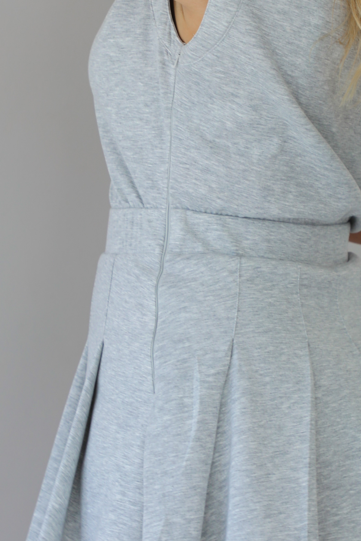 A woman modeling a light gray heathered short-sleeve collared dress with a pleated mini skirt, indoors against a white wall. The photo shows a closeup of the zipper closure on the side of the dress. 