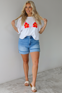 Woman wearing a white cropped boxy tee with a pink, red, and yellow bikini graphic on the front with a pair of medium wash denim shorts indoors against a white wall with white marble flooring.