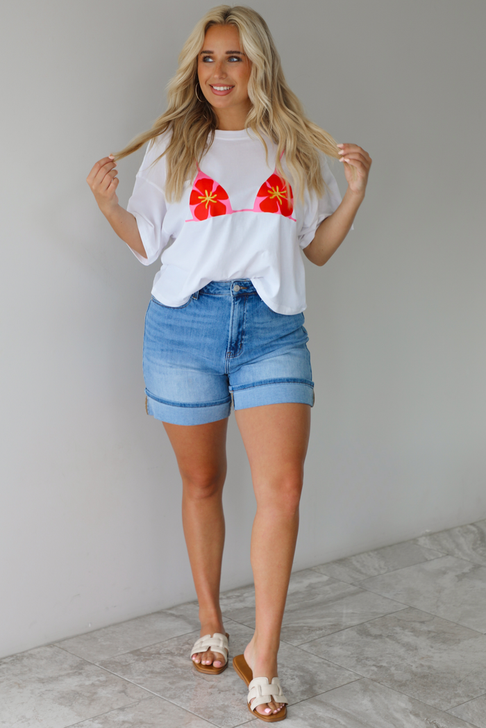 Woman wearing a white cropped boxy tee with a pink, red, and yellow bikini graphic on the front with a pair of medium wash denim shorts indoors against a white wall with white marble flooring.