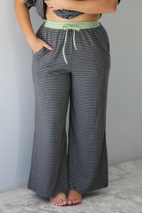 A woman shown from the waist down wearing a charcoal and white striped two piece matching set with green trim detailing at the neckline and waistline, featuring an oversized short sleeved top and high waisted pants, standing indoors against a white wall with white marble flooring. The photo shows a closeup of the pants with the models hand in one of the side pockets. 