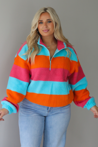 A woman wearing a turquoise, pink, and orange striped sherpa quarter-zip sweater with light wash denim jeans, standing indoors against a white wall.