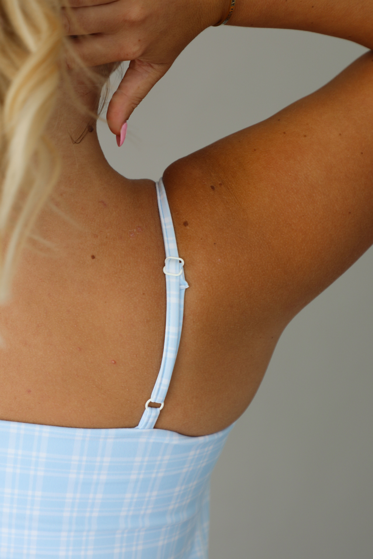 A woman wearing a light blue and white plaid mini dress, standing indoors against a white wall. The photo shows a closeup of the adjustable straps. 