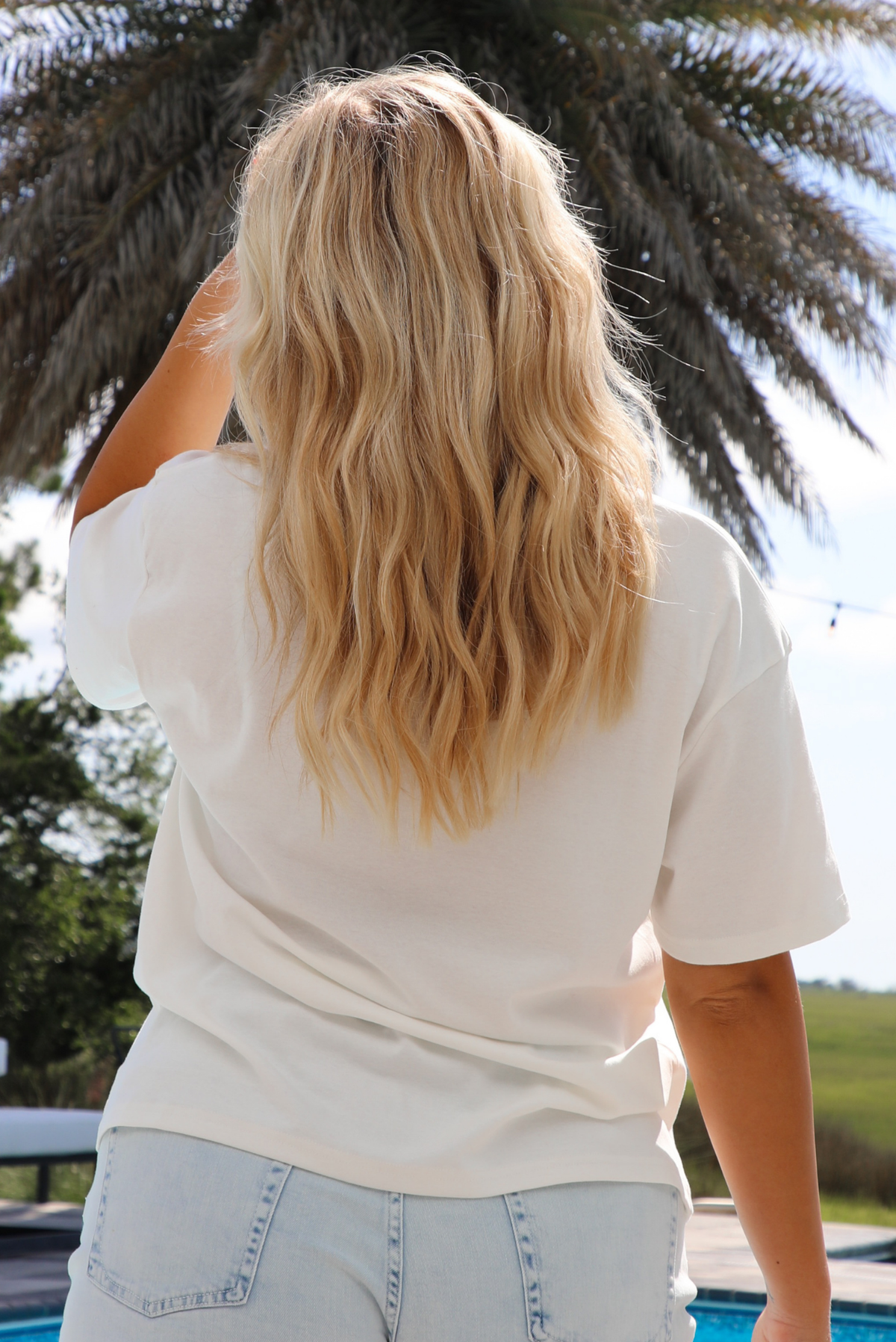 Woman wearing a white tee with a navy graphic on the front of a swimsuit top with light wash denim shorts front of a pool and palm tree in the background. Th photo shows the back of the tee. 
