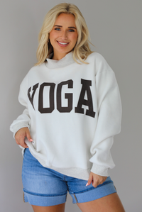 Woman wearing a heather grey oversized sweatshirt with the word 'PILATES' written in collegiate font on the front with medium wash denim shorts indoors against a white wall.