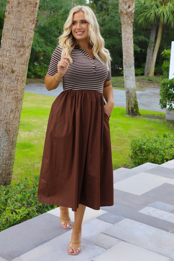 Woman model wearing a brown midi length dress that has a brown and white striped bodice, and brown midi length skirt. The photo is taken outdoors with greenery in the back.