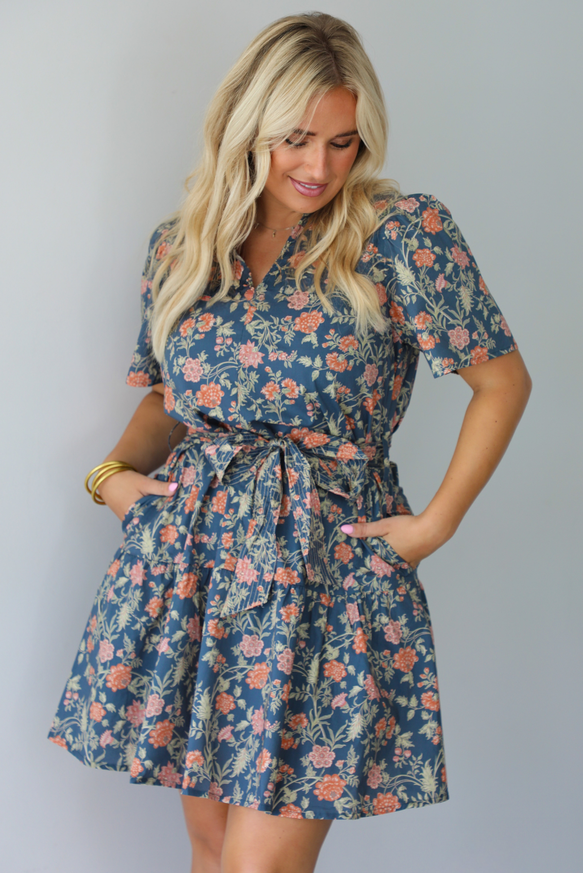 A woman modeling a short-sleeve floral mini dress with a tie waist, standing indoors against a white wall.