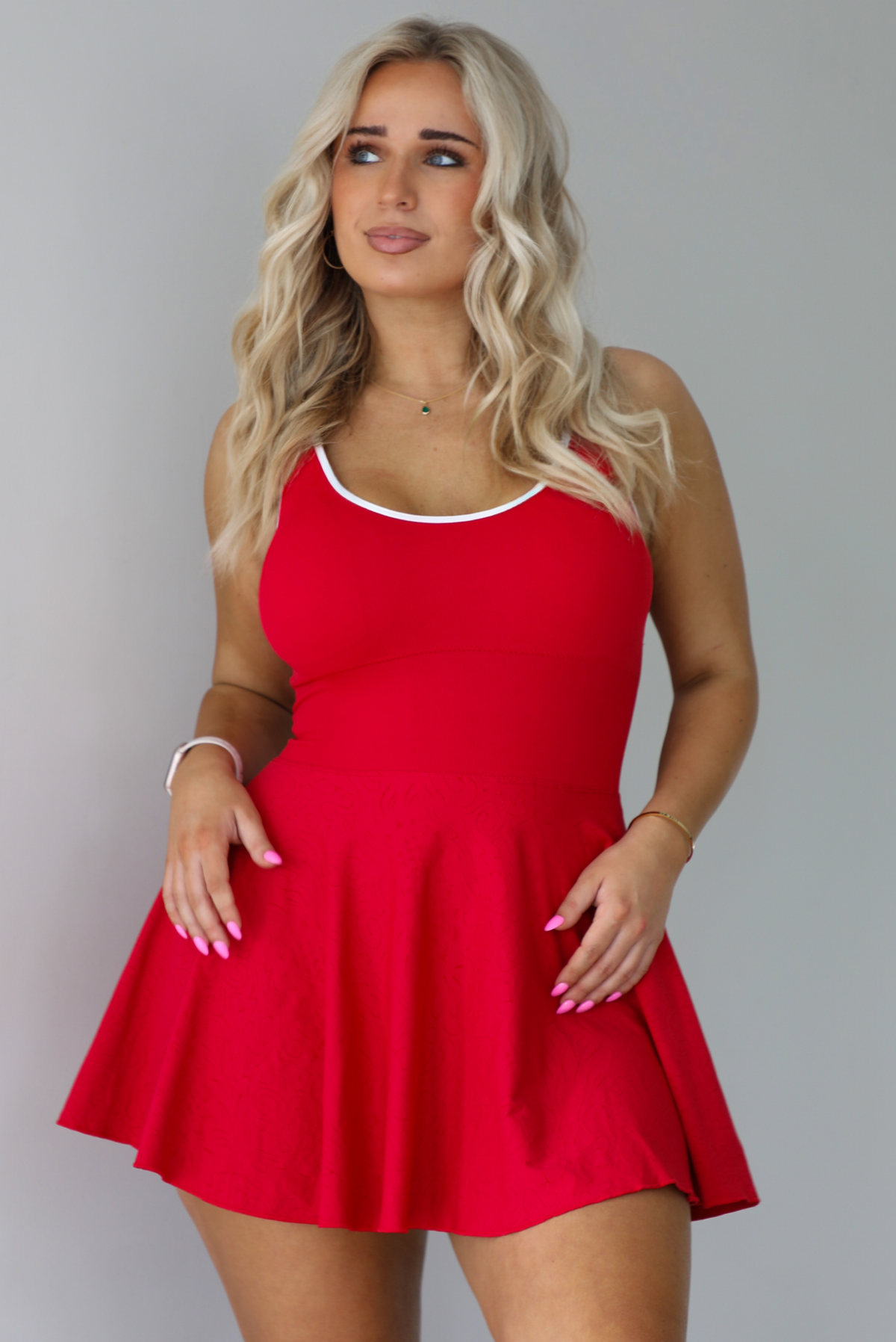 Woman wearing a red athleisure dress that has white trim at the top of the bodice, a fitted bodice, and flowy skirt. The photo is taken indoors against a white wall.