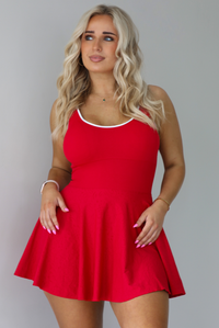 Woman wearing a red athleisure dress that has white trim at the top of the bodice, a fitted bodice, and flowy skirt. The photo is taken indoors against a white wall.