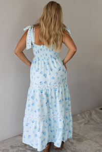 Woman wearing a light blue floral maxi dress indoors against a white wall with white marble flooring. The photo shows the back of the dress, which features a smocked detail at the back.
