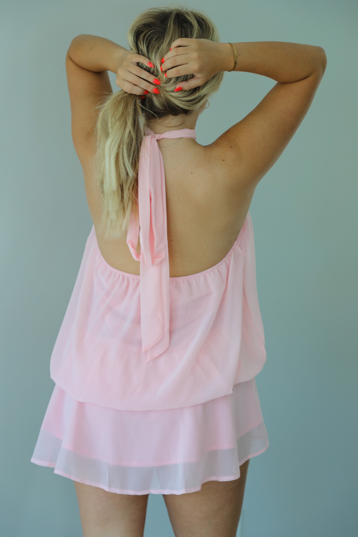 Woman wearing a pink dress that has a halter tie neckline, a cinched detail at the hips, and a flowy design indoors against a white wall. The photo shows the back of the dress, which has an open back and halter tie neckline. 