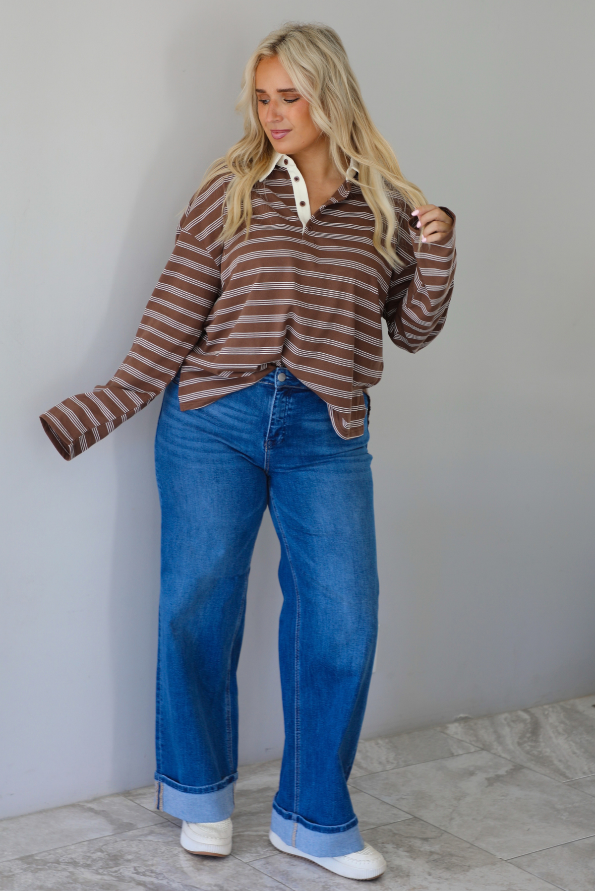 A woman wearing a brown and cream striped collared quarter-button long sleeved top, front tucked into dark wash denim wide leg jeans that have a cuffed hem, paired with cream sneakers, standing indoors against a white wall with white marble flooring. 