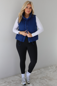 Woman wearing a navy colored puffer vest, white long-sleeve shirt, black leggings, and gray sneakers with white ankle socks indoors against a white wall with white marble flooring.