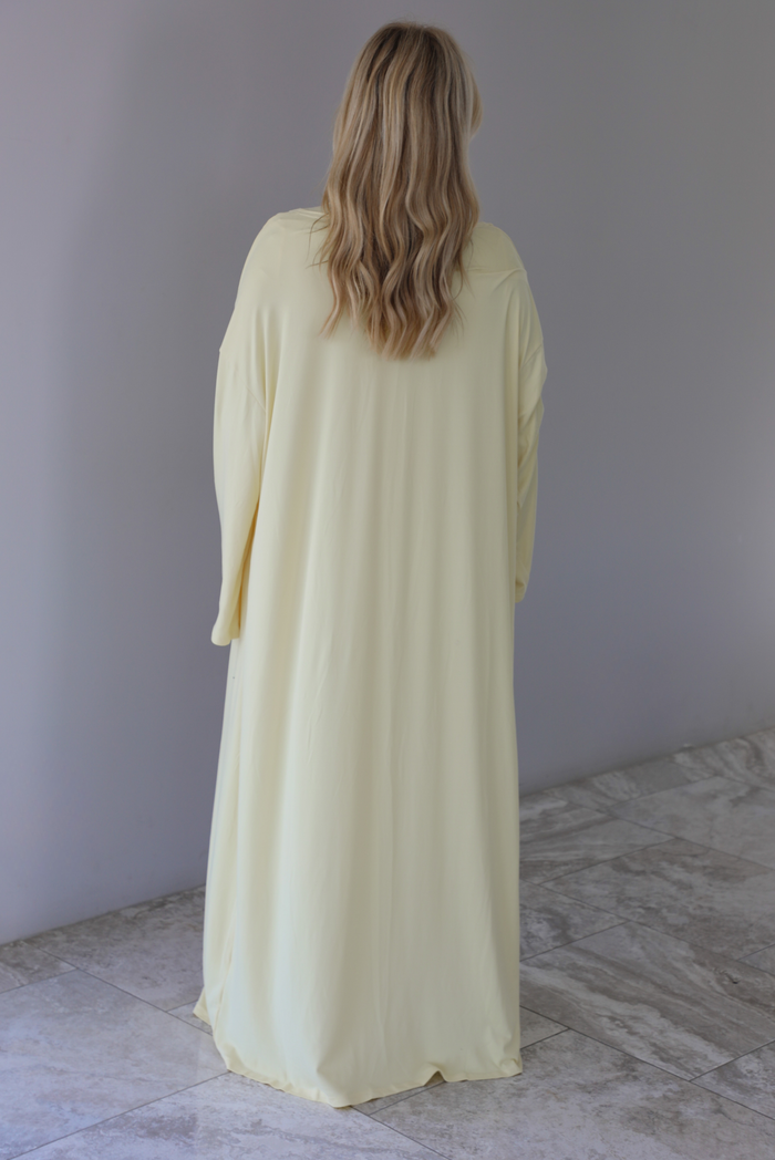 Woman wearing a light yellow three piece set featuring a long sleeved top, long, flared hem pants, and a long, matching cardigan. The photo is taken indoors against a white wall with white marble flooring. The photo shows the back of the cardigan. 