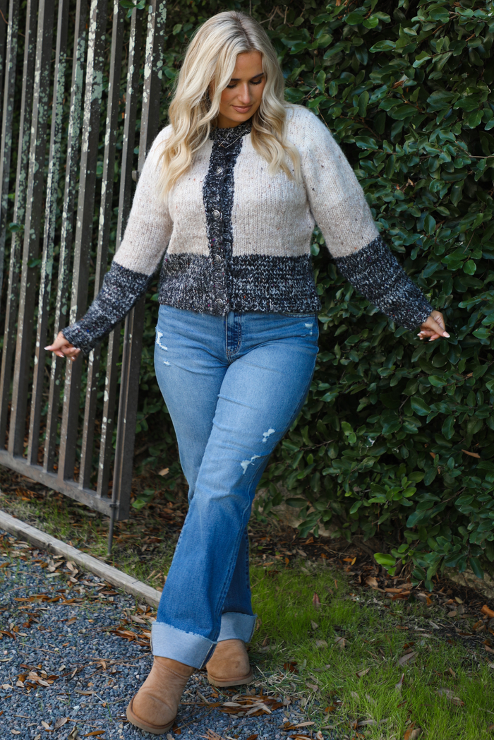 Colorblock designed cardigan with ivory, sand, oatmeal, and charcoal colors. The cardigan has functional button closures in the front with a relaxed fit. The cardigan is styled with medium wash denim. The photo is taken outdoors with greenery in the back. 