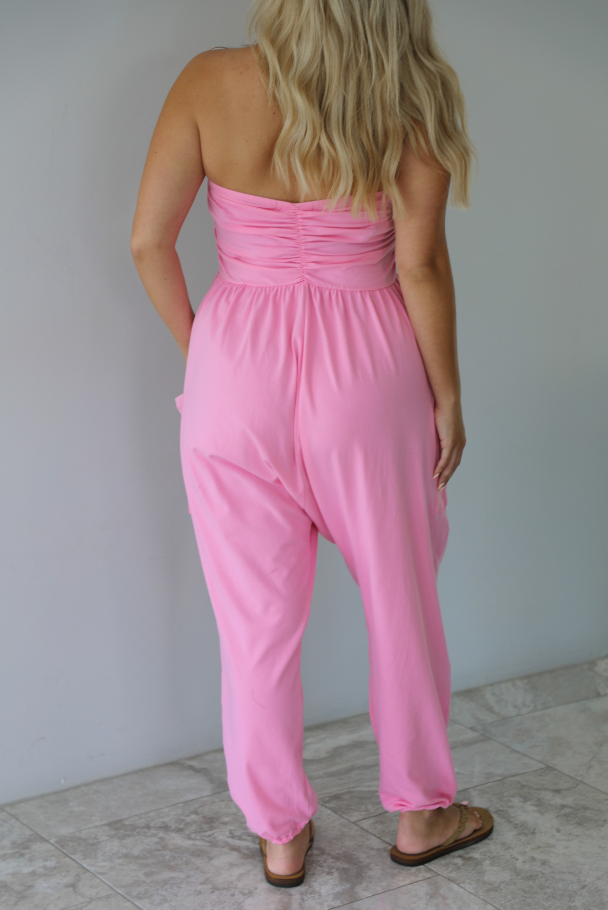 Woman wearing a pink strapless jumpsuit with a fitted bodice and slouchy pants indoors against a white wall with white marble flooring. The photo shows the back of the jumpsuit. 