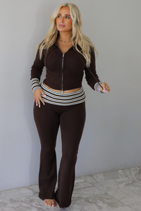 A woman modeling a dark brown ribbed zip-up hoodie and matching flared pants set with white striped accents on the cuffs and waistband, standing indoors against a white wall with white marble flooring. 