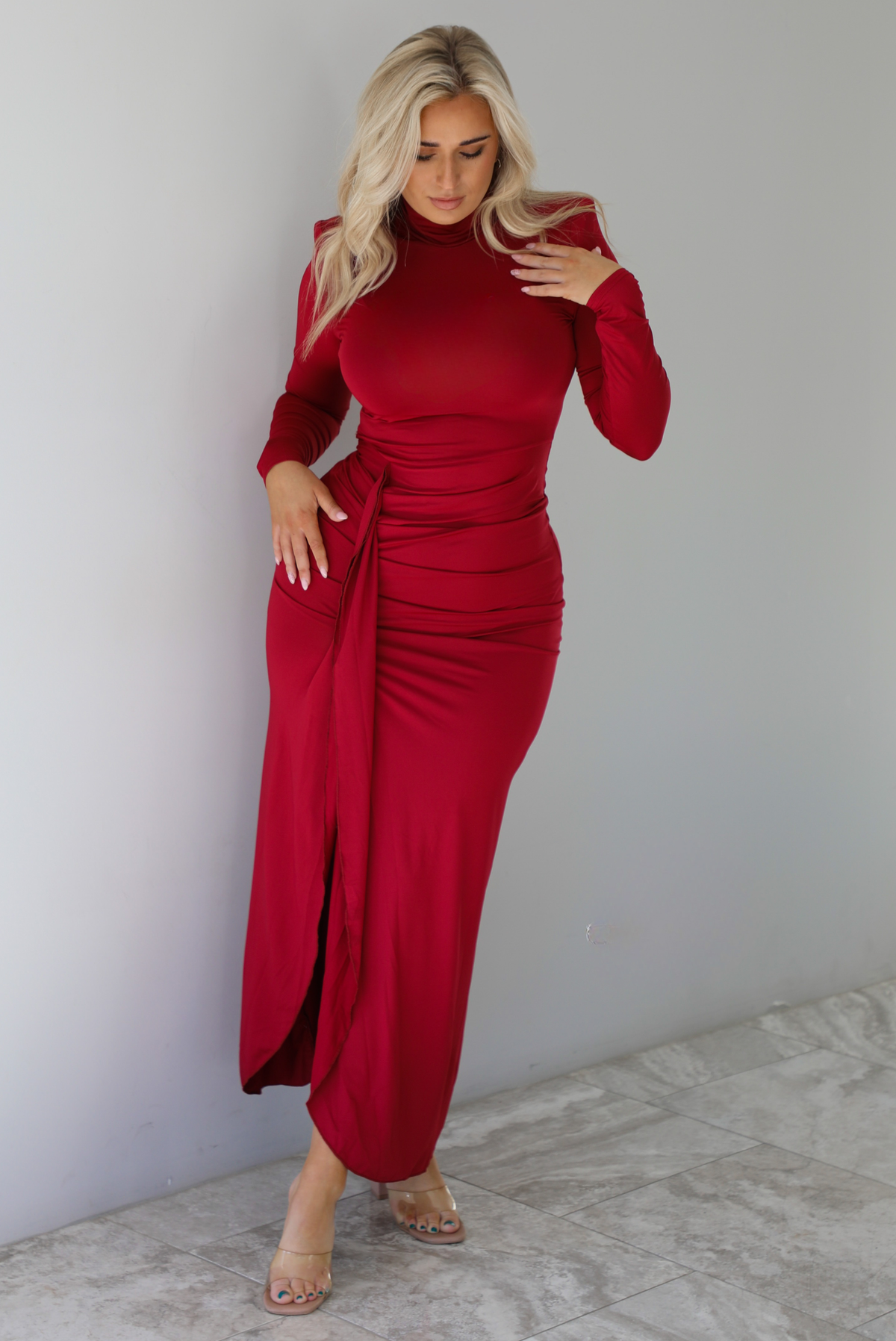 Woman wearing a red maxi dress that has a high neckline, shoulder pads, a fitted bodice, long sleeves, with a split hem detail on the left side. The photo is taken indoors against a white wall with white marble flooring. 