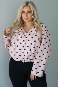 A woman wearing a pink button-down jacket with a black polka dot print pattern, paired with black leggings, white ankle socks, and black slide on slippers, standing indoors against a white wall.